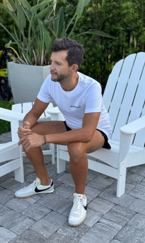 Man sitting wearing Unrefined Friends white pride shirt with rainbow logo outdoors.