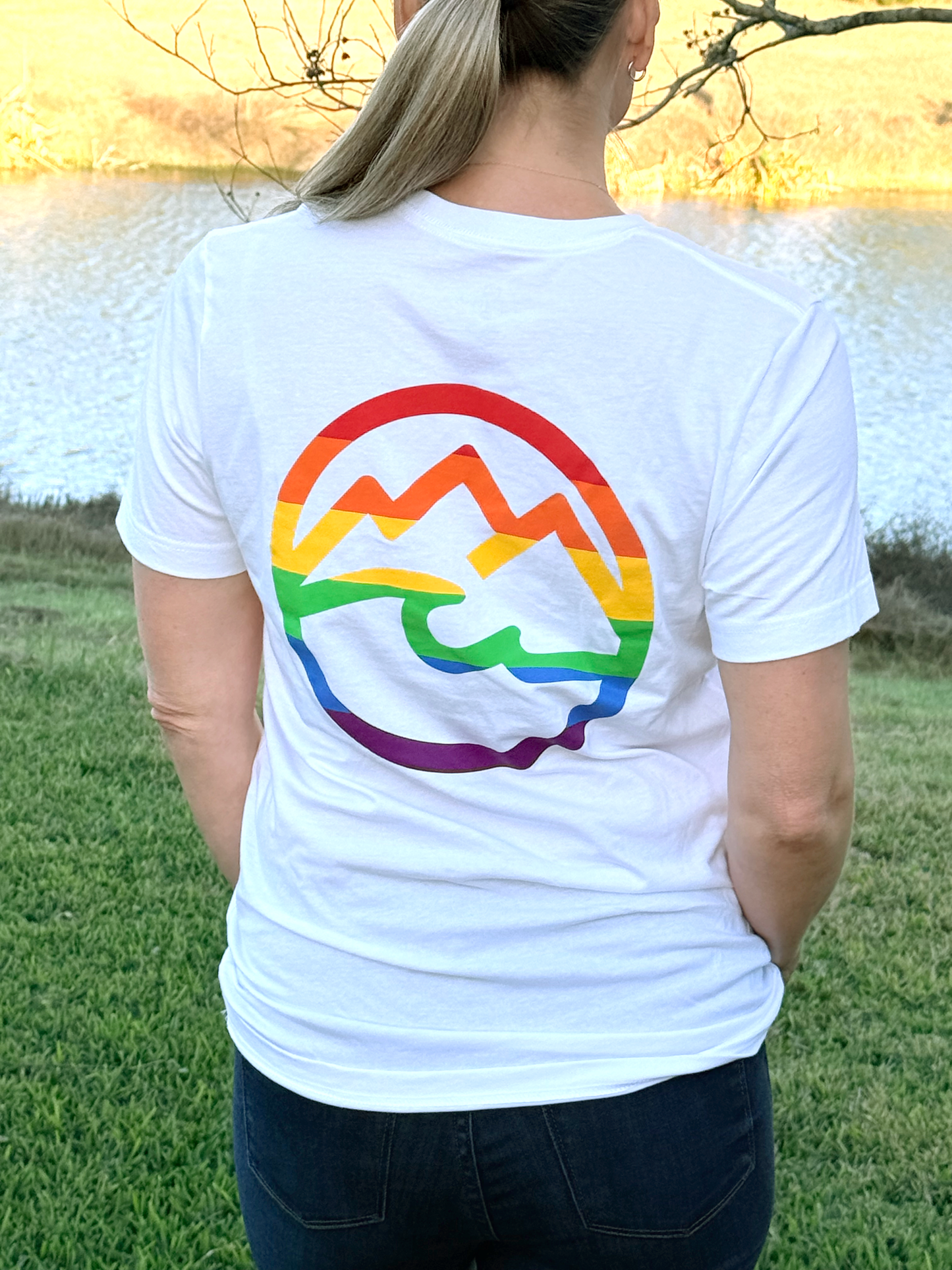 Woman facing away wearing a white t-shirt with a rainbow mountain and ocean design outdoors.