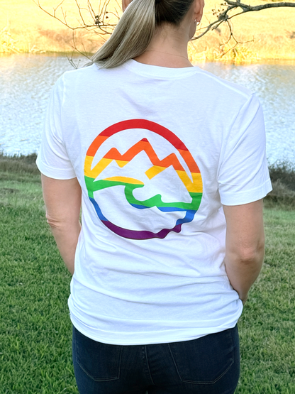 Woman facing away wearing a white t-shirt with a rainbow mountain and ocean design outdoors.
