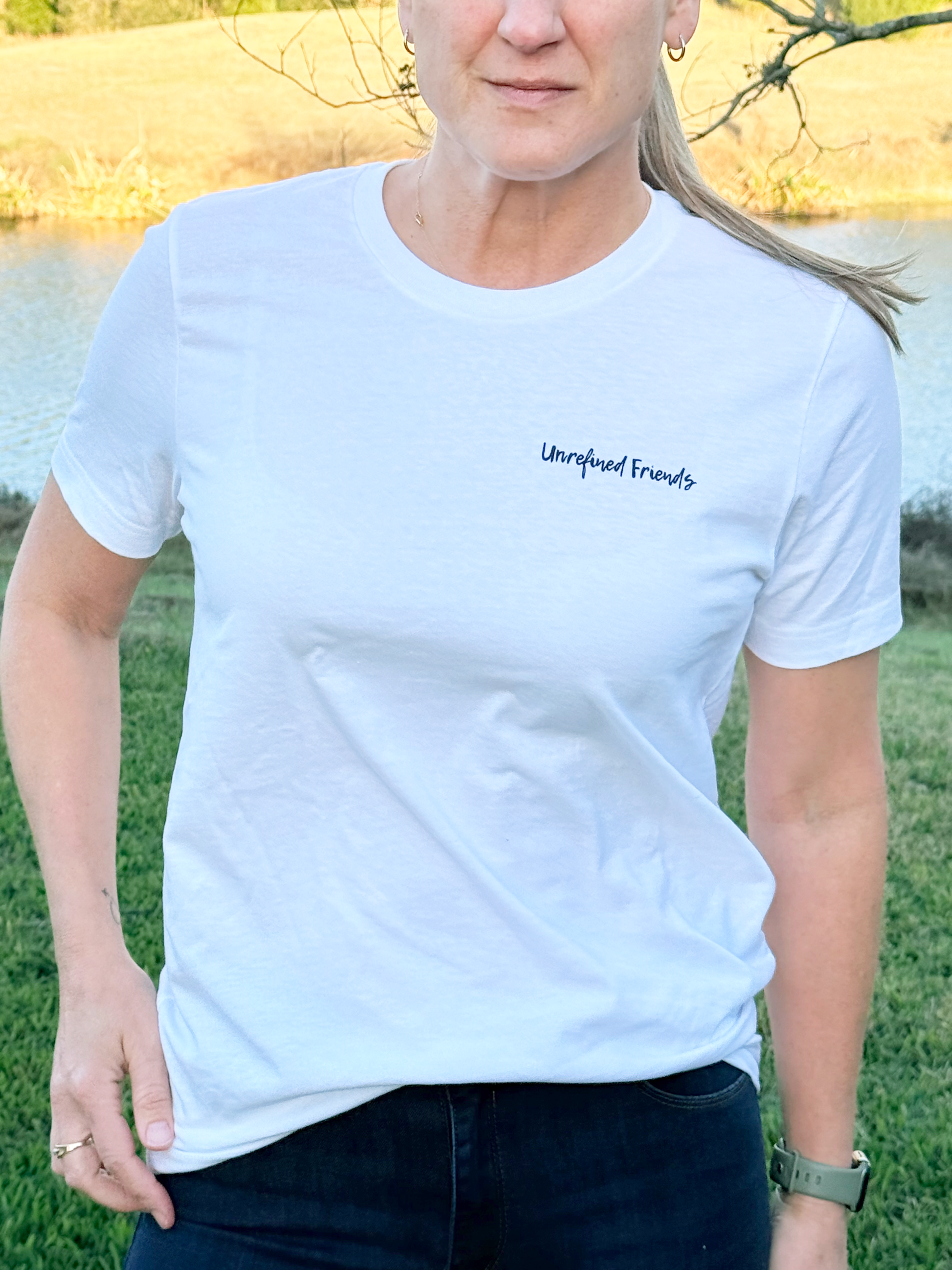 Person wearing a white t-shirt with purple script 'Unrefined Friends' text outdoors.